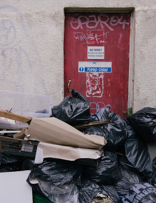 A pile of assorted household rubbish, including cardboard boxes, paper bags, and plastic wrapping, is accumulated against a weathered brick wall and a concrete block wall in an outdoor urban setting.