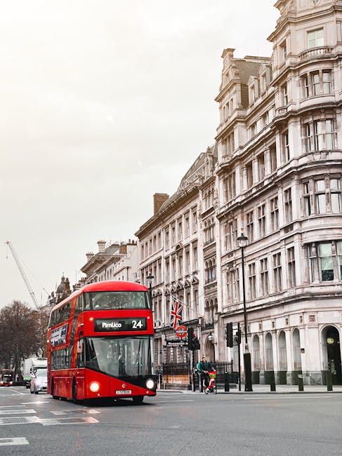 A red double-decker bus traveling along a street in an urban area, with large, ornate white stone buildings featuring multiple windows and architectural detailing in the background. The bus displays t